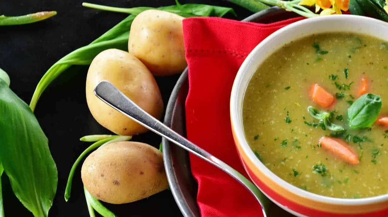 Three spuds and vegetable leaves next to a bowl of delicious potato and veggie soup, garnished with basil.